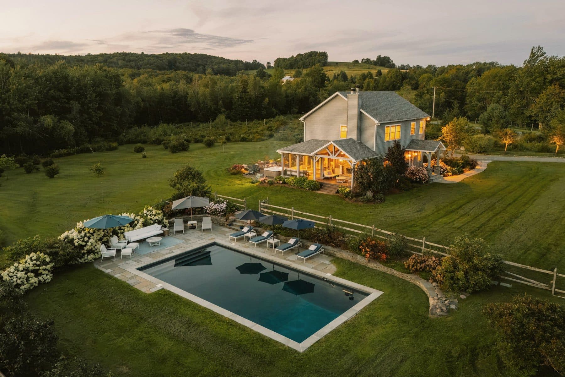 Aerial view at dusk of a country house extended with new wings, a covered porch and a pool terrace.